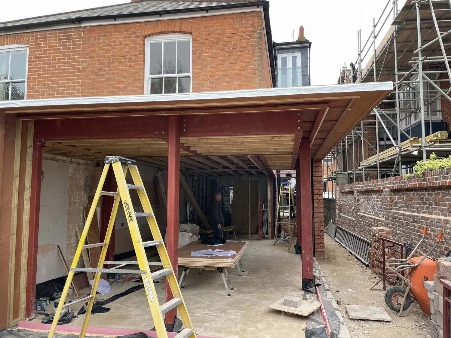 Construction site where a building extension is being added to a brick house, featuring scaffolding, ladders, wooden framework, and construction workers.