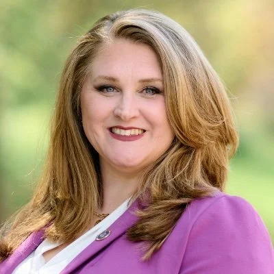 A woman with shoulder-length blonde hair smiling outdoors, wearing a purple blazer and white blouse.
