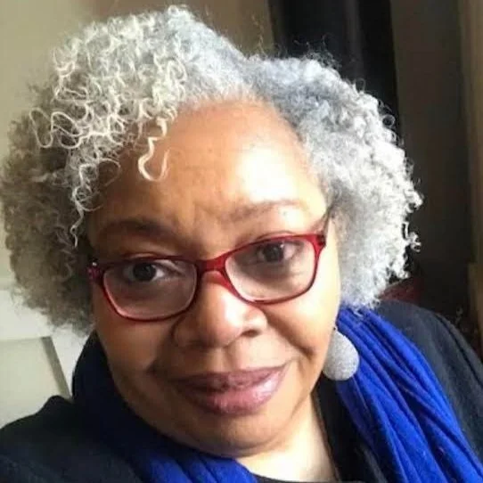 A woman with curly gray and white hair, wearing red glasses and a blue scarf, smiling at the camera.