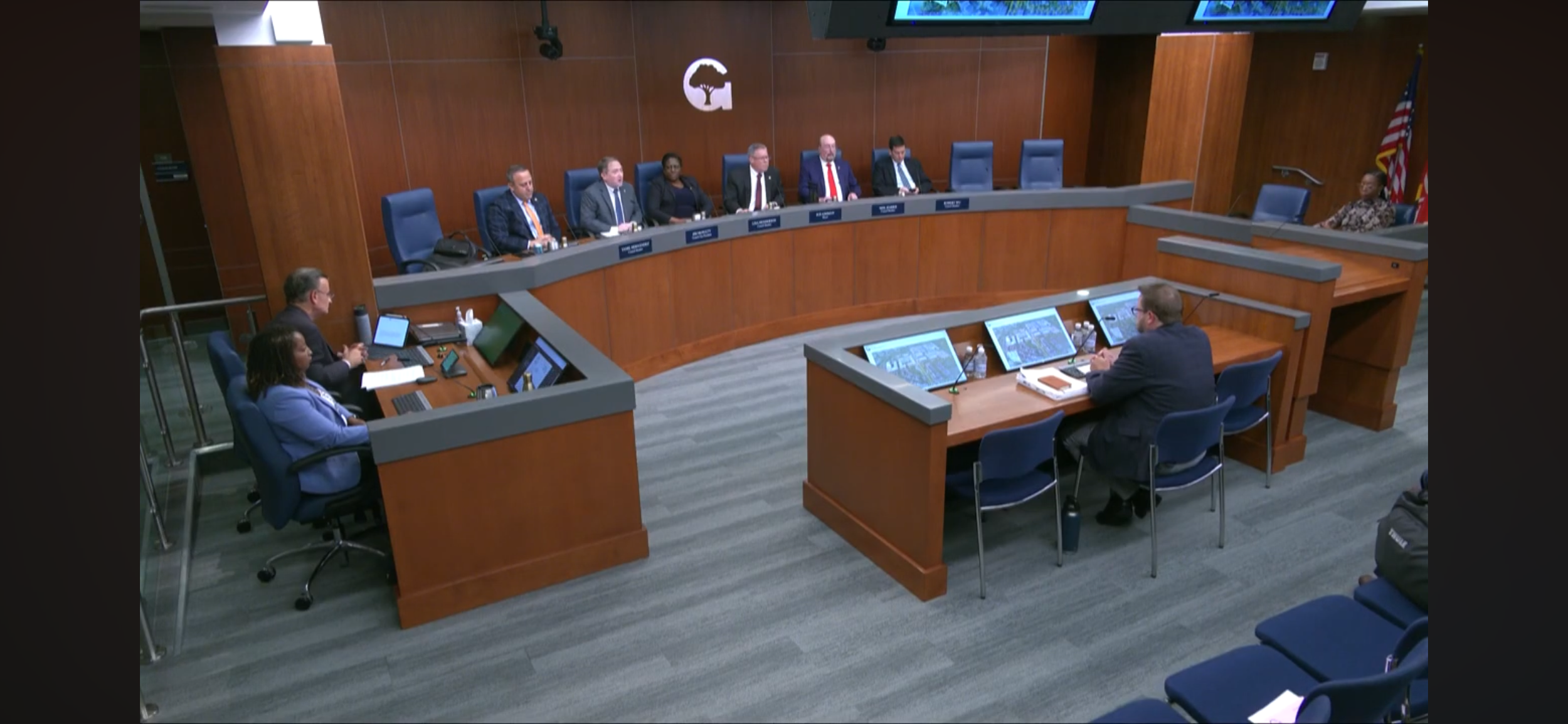 A formal city council or governmental meeting room with panel of officials seated at a curved wooden desk and an audience area. Four people are visible at a separate table in the foreground with electronic devices and documents. The room features wood-paneled walls with an emblem and American flags.