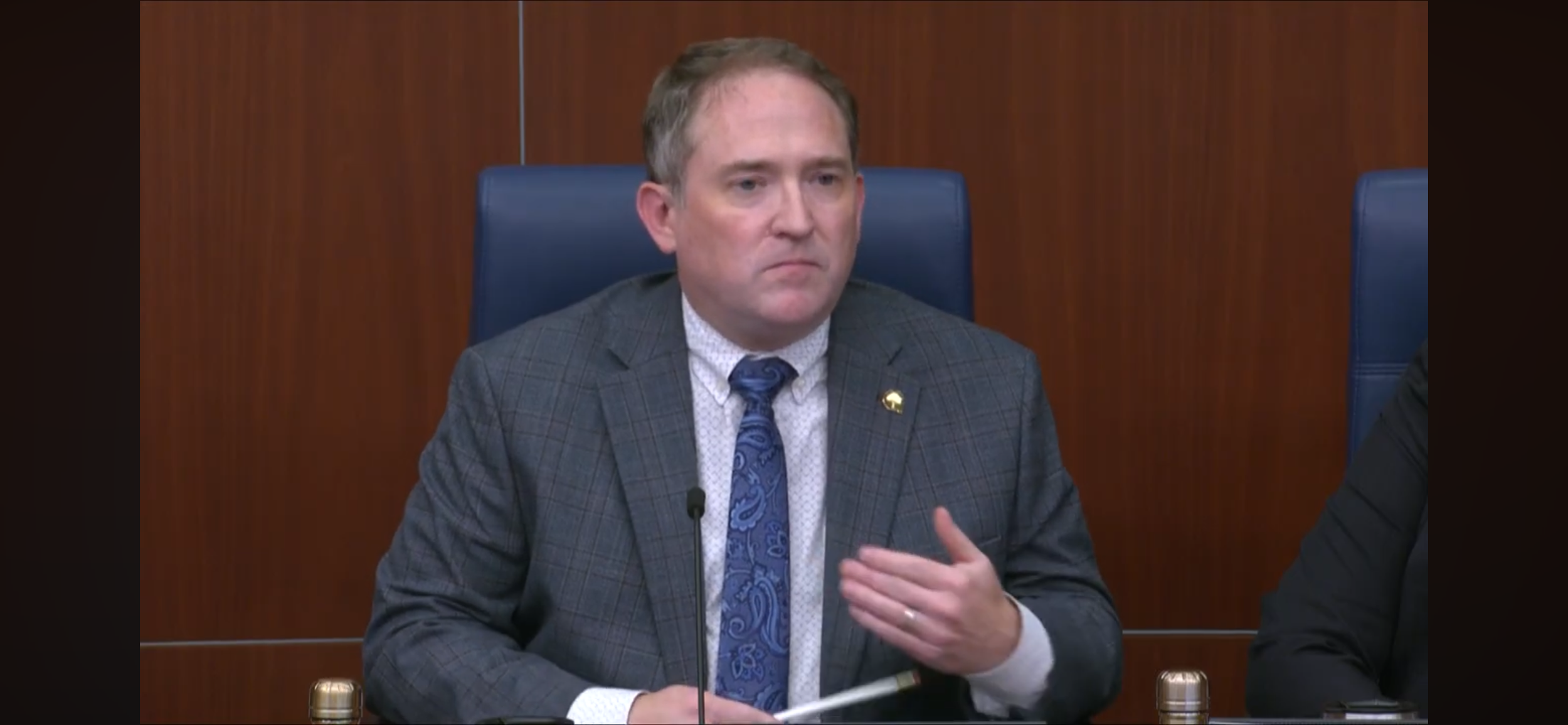 Council Member Mculty in a gray plaid suit, wearing a blue paisley tie, sitting at a table, speaking into a microphone during a formal meeting.