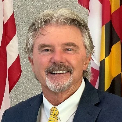 A smiling man with gray hair and a beard, wearing a dark suit, white shirt, and yellow tie, standing in front of American and Maryland flags.