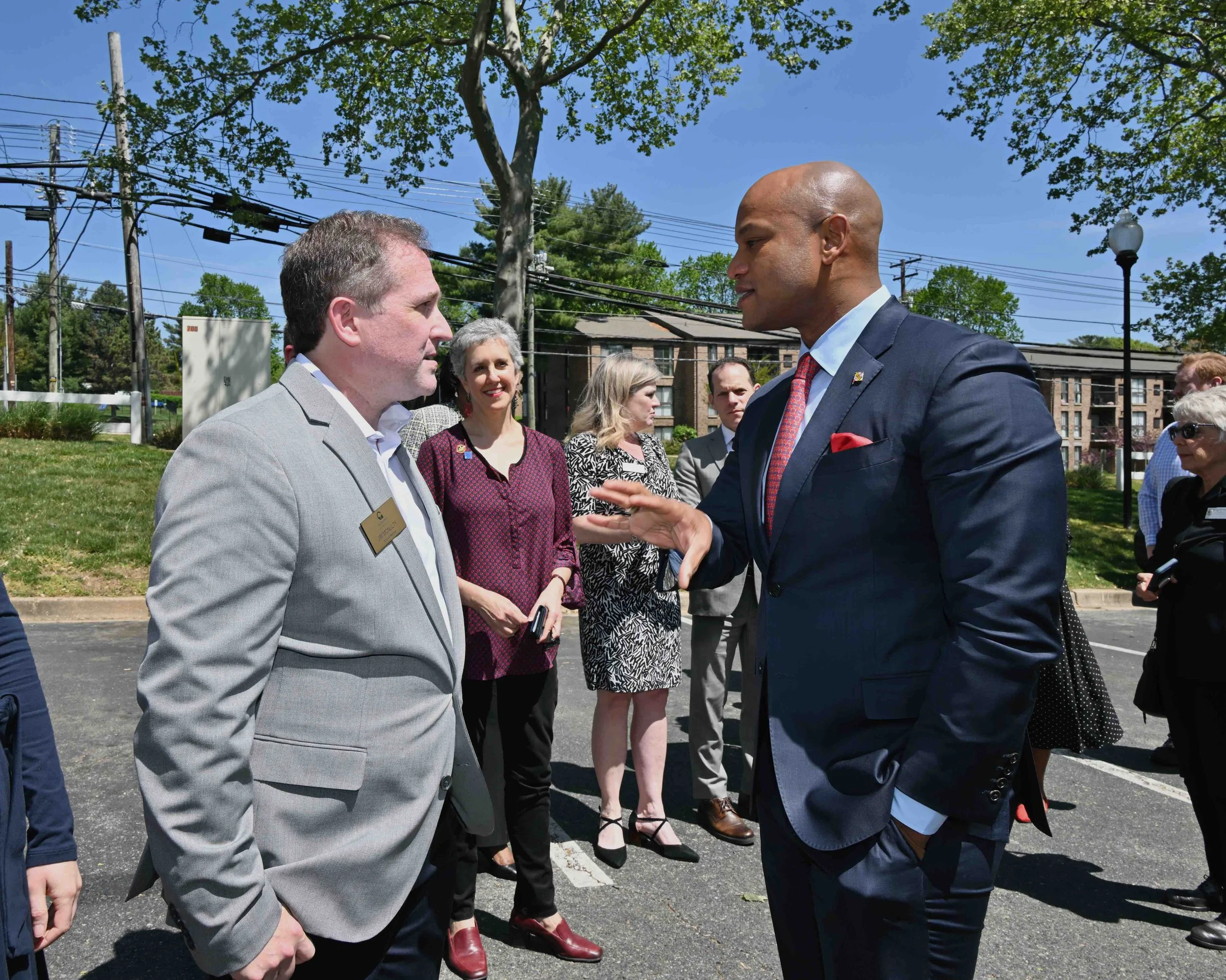 Two men are having a conversation outdoors on a sunny day, with several people in the background, some holding phones, and trees, power lines, and buildings in the distance.