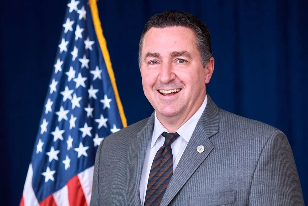 Smiling man in gray suit with a striped tie standing in front of an American flag