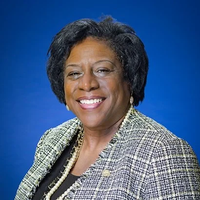 Portrait of Council Member Lisa Henderson, a woman with short black hair, wearing a plaid blazer, pearl earrings, and a pearl necklace, smiling against a blue background.