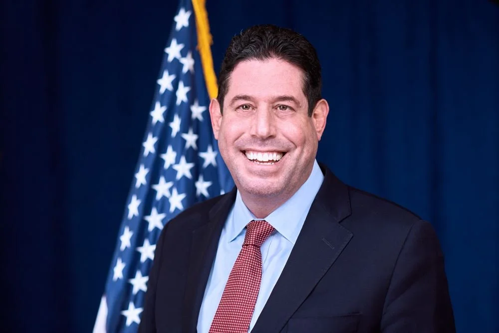 A man in a business suit smiling in front of an American flag.