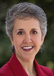 Headshot of Senator Cheryl Kagan, a woman with short gray hair smiling, wearing a red blazer and earrings, outdoors with greenery in the background.