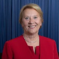 Portrait of a smiling woman with blonde hair wearing a red blazer against a blue background.