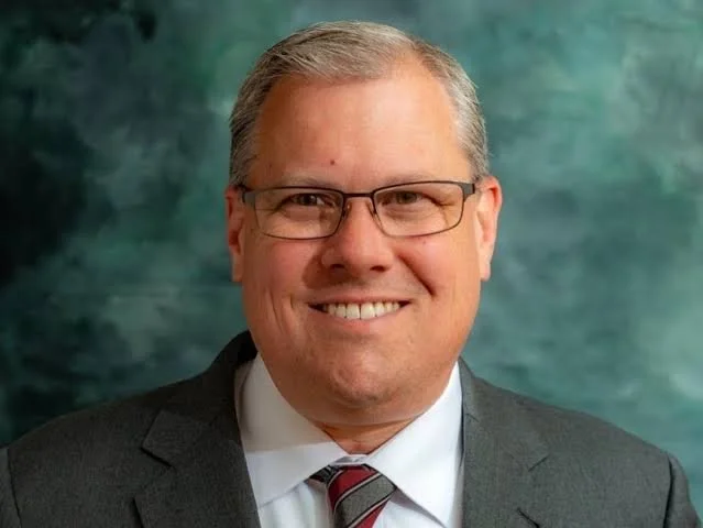 Headshot of Jud Ashman, a smiling man with gray hair, wearing glasses, a dark suit, white dress shirt, and red-striped tie, against a greenish abstract background.