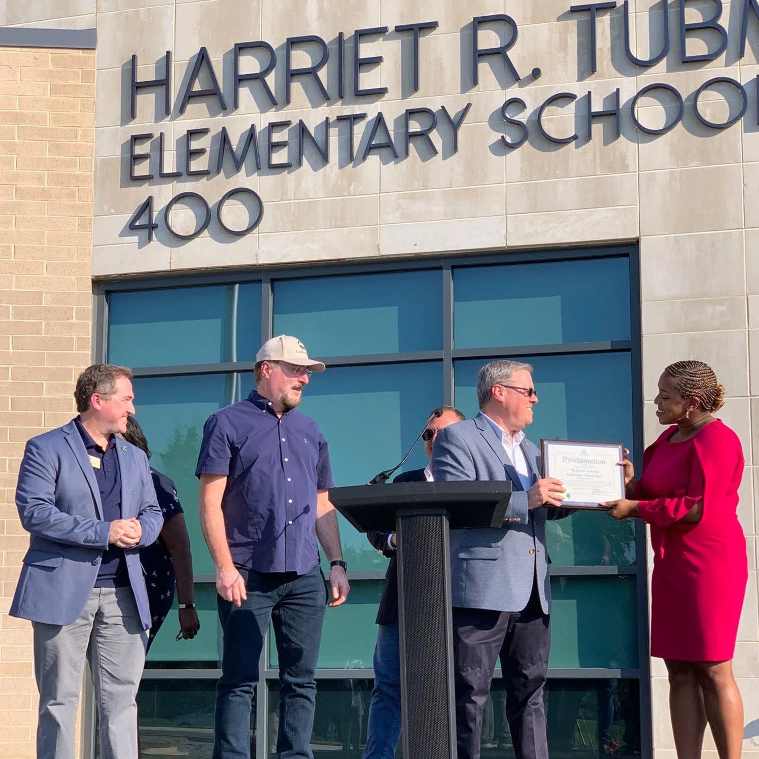 A woman in a pink dress is presenting a proclamation certificate to a man in a gray suit outside Harriet R. TUB Elementary School at 400. Four other people are standing nearby, and a podium is visible in the scene.