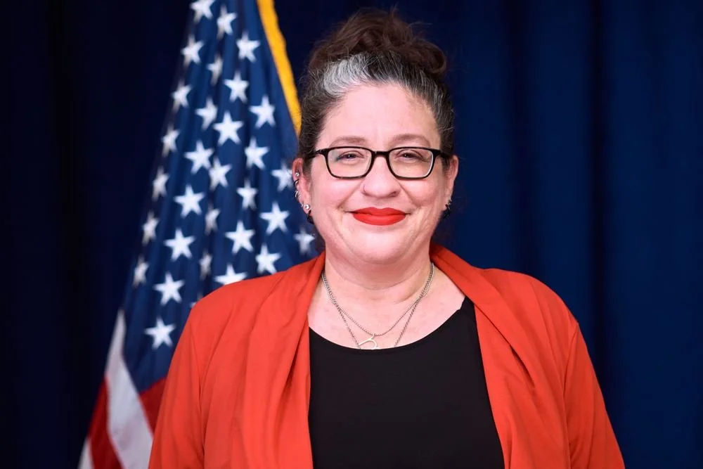 A woman with glasses, wearing a red jacket and a black top, standing in front of an American flag with a dark blue background.