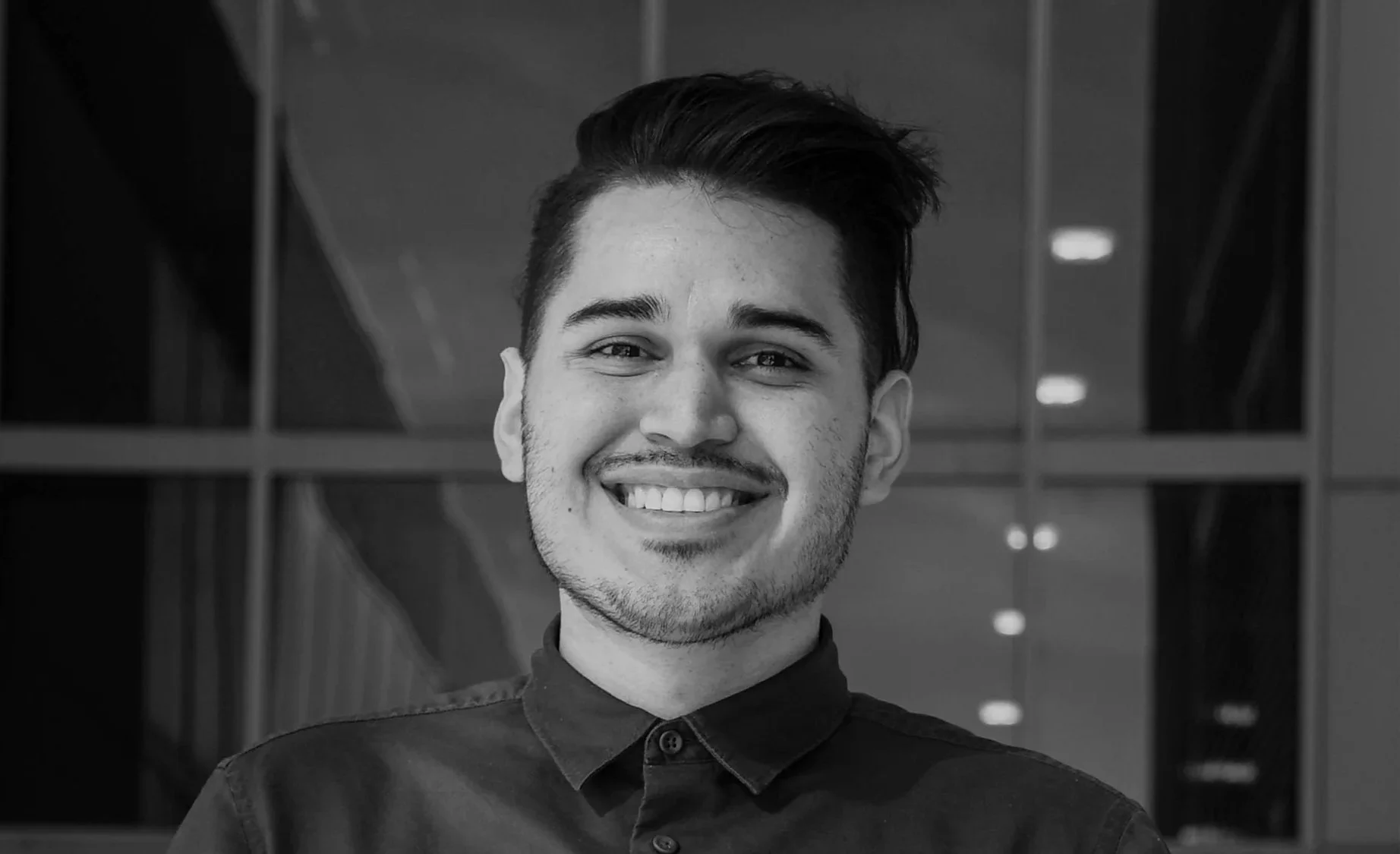 Black and white photo of a smiling young man with short dark hair, wearing a collared shirt, standing indoors with a modern architectural background.
