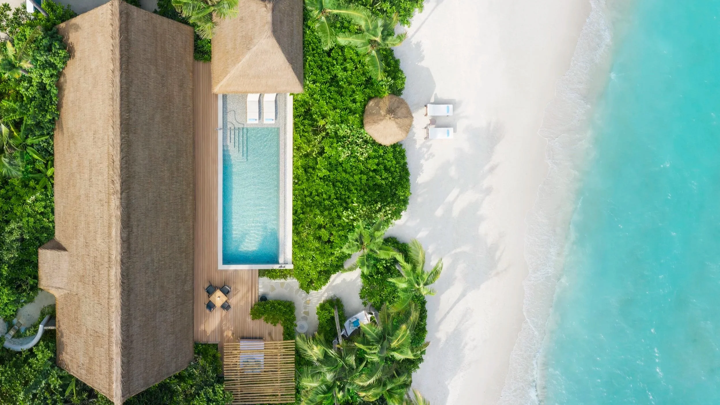 Aerial view of a beach villa with private pool, beach umbrella and two deck chairs on sand next to lagoon