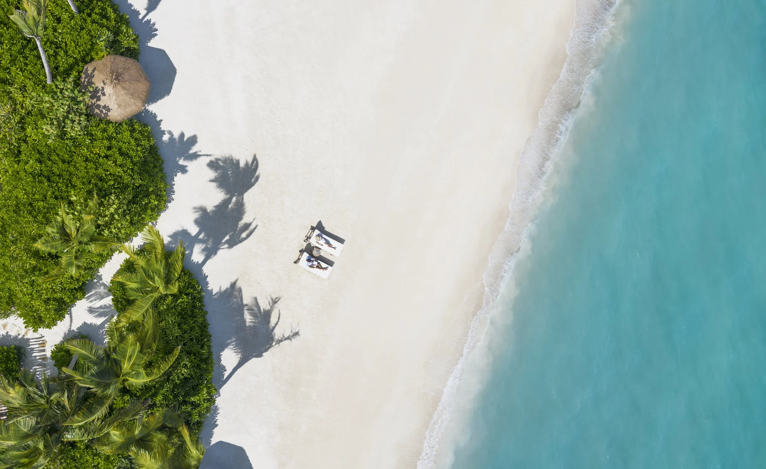 Two people lying on deck chairs on a white sand beach and turquoise lagoon