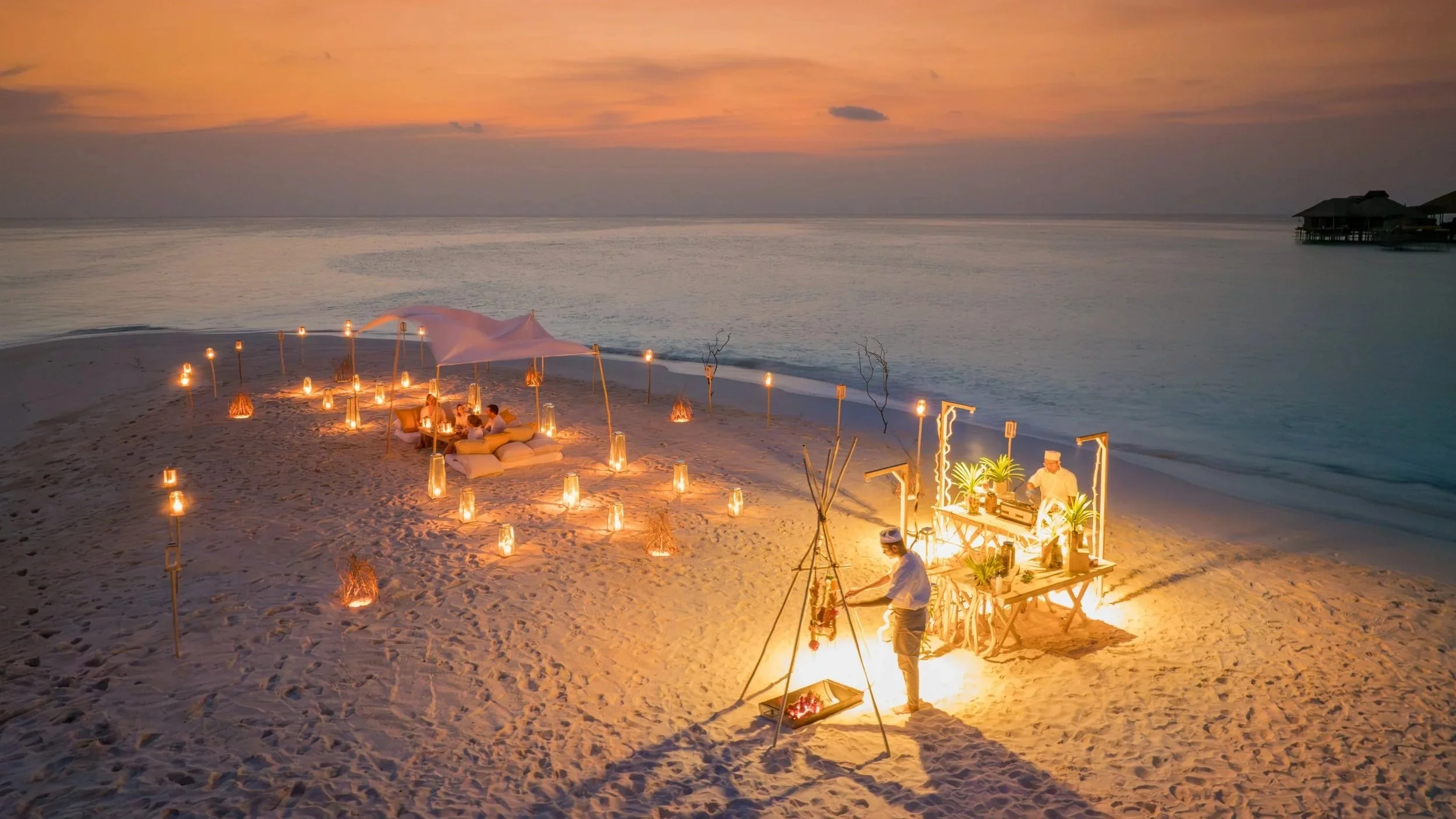 sunset on a sandbank with chef making a bbq dinner and couple  relaxing on cushions surrounded by lights