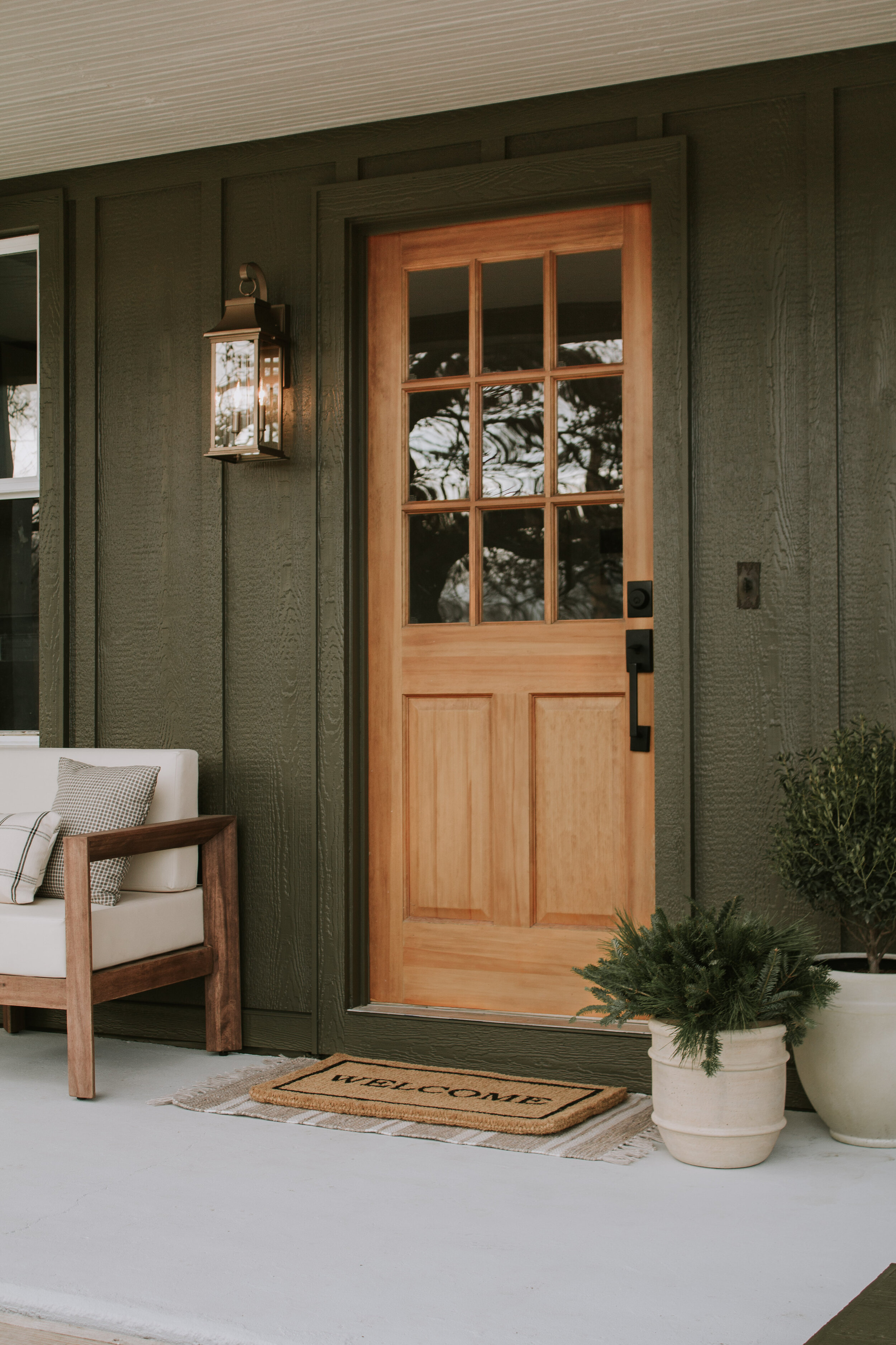A closeup of our front entry porch & 15 layered entry rug combos - Nadine Stay | Outdoor club chairs with a wood frame and ivory cushions, wood front door with window panes, doormat, aged outdoor pots, and an antique outdoor lantern sconce. Cabin po…