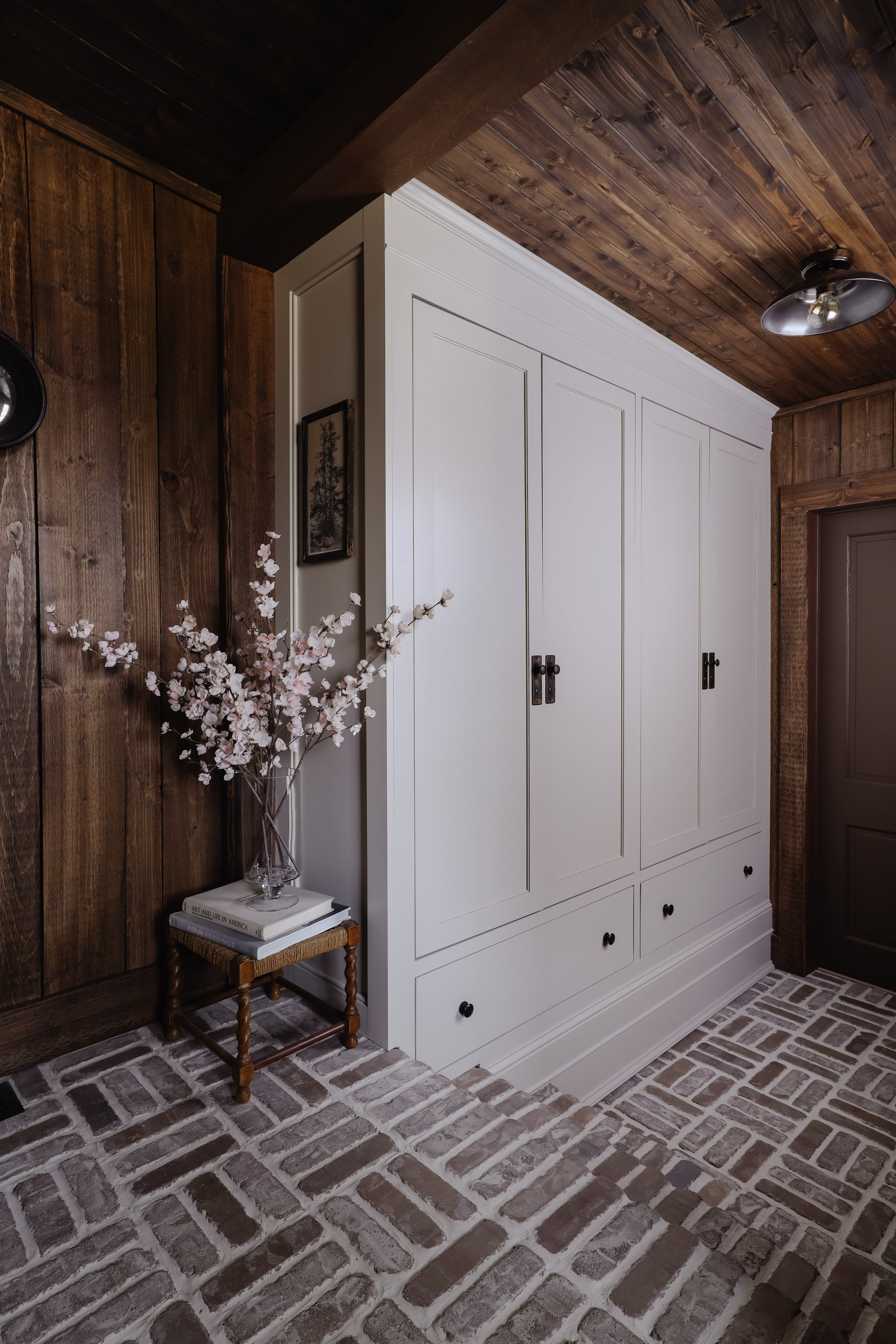 Rustic moody laundry room and mudroom. Dark wood walls. Basketweave brick floors in laundry room. Beige paint color for cabinets and brown paint color for doors. Built in closet for mud room. Nadine Stay