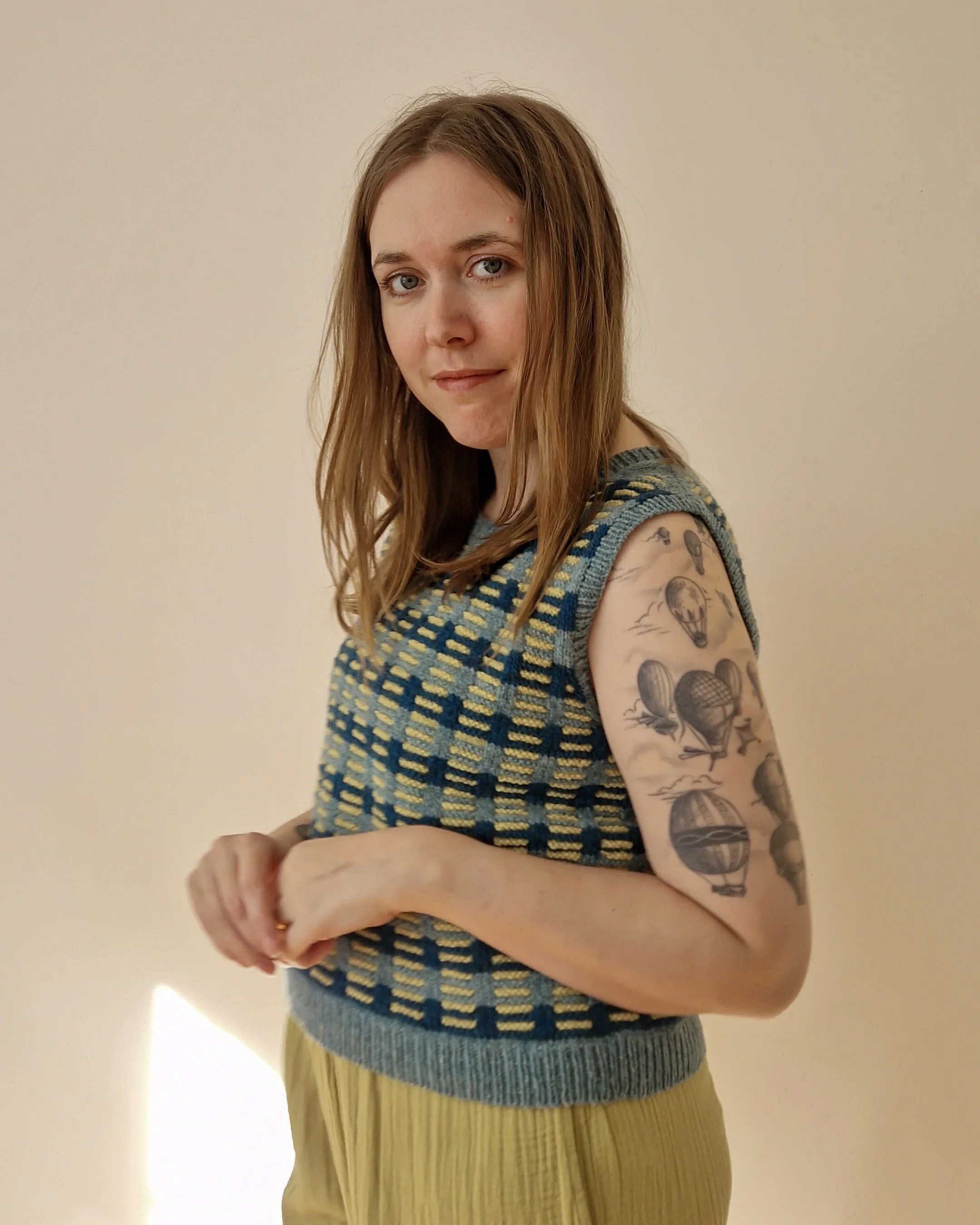 Clare is wearing a handknitted slipover in blues and yellow, and slightly smiling in side profile at the camers