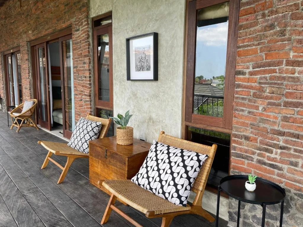 Outdoor balcony with wooden flooring, brick and stucco walls, glass doors, and seating including two wooden chairs with black and white patterned cushions, a small wooden table with a potted plant, and a black side table with a small potted plant.