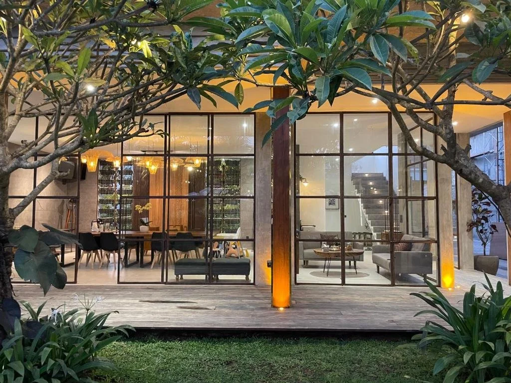 Front view of a modern house with a large glass wall, visible interior living and dining areas, surrounded by green plants and trees, with a wooden deck outside.