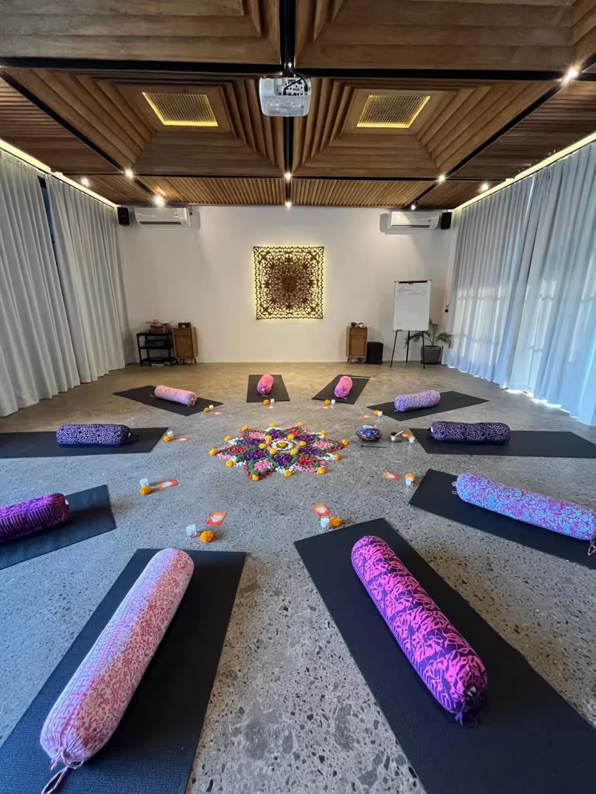 A yoga studio set up for a meditation or yoga class with eight black mats, each with pink and purple bolsters, arranged in a circle on a terrazzo floor. In the center, there is a colorful floral arrangement. Small candles or decorative items are plac