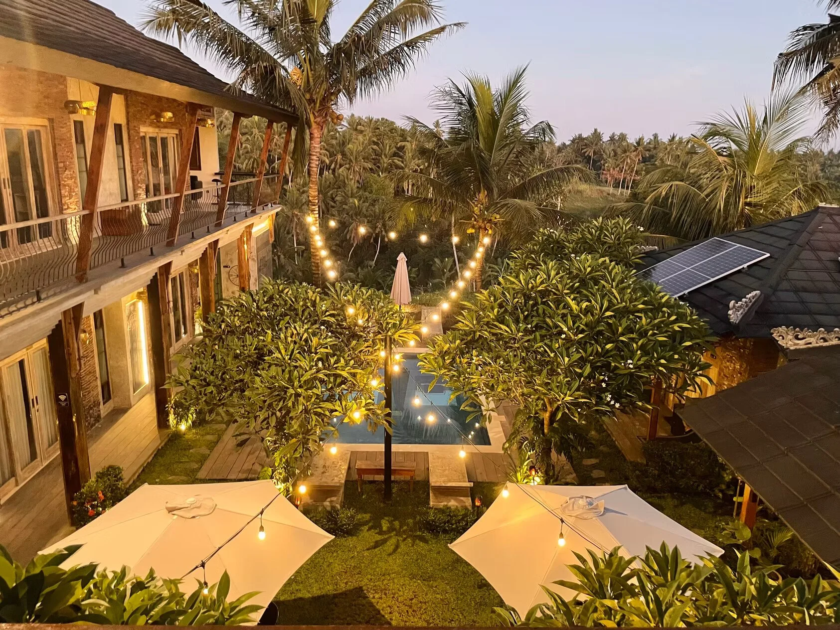 A backyard scene at dusk with a swimming pool, shaded by large trees, with string lights hanging between the trees. There are two large umbrellas with lights underneath, surrounding the lawn area, and a building with a balcony visible on the left sid