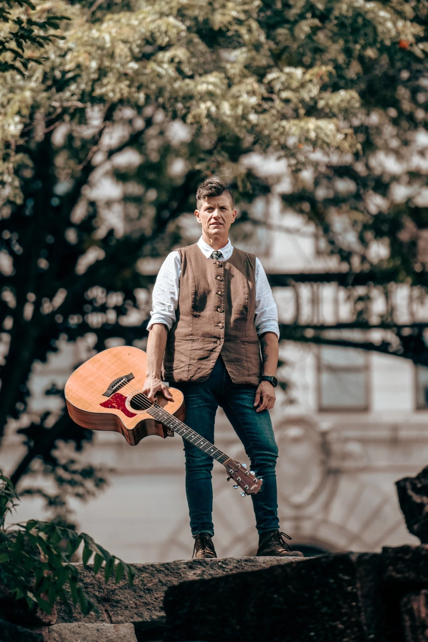 Andrea Paquin posing for photo holding guitar in hand standing on top of stone wall with trees in the background