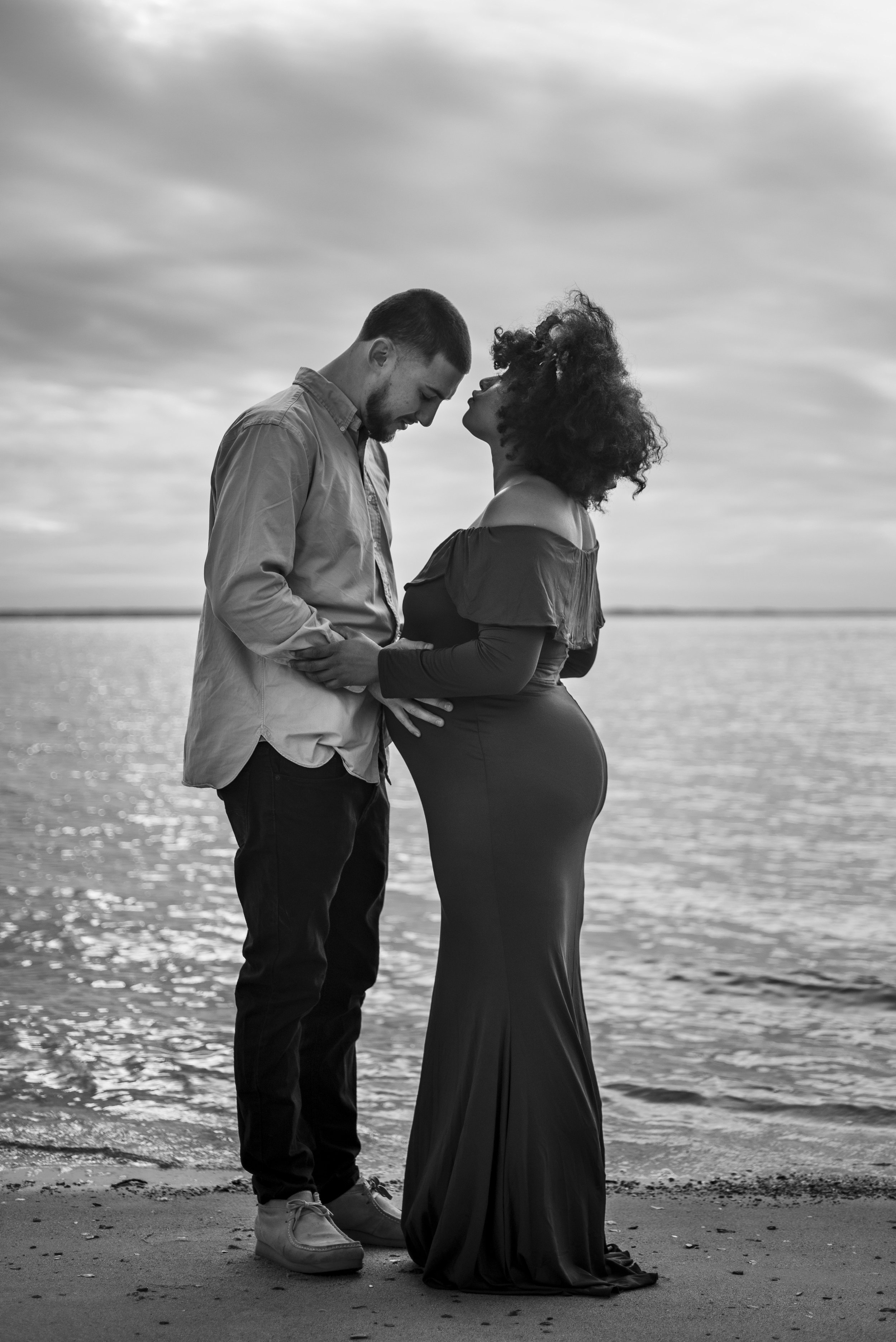 woman kiss mans forehead on the beach by the shore as they embrace her baby bump.