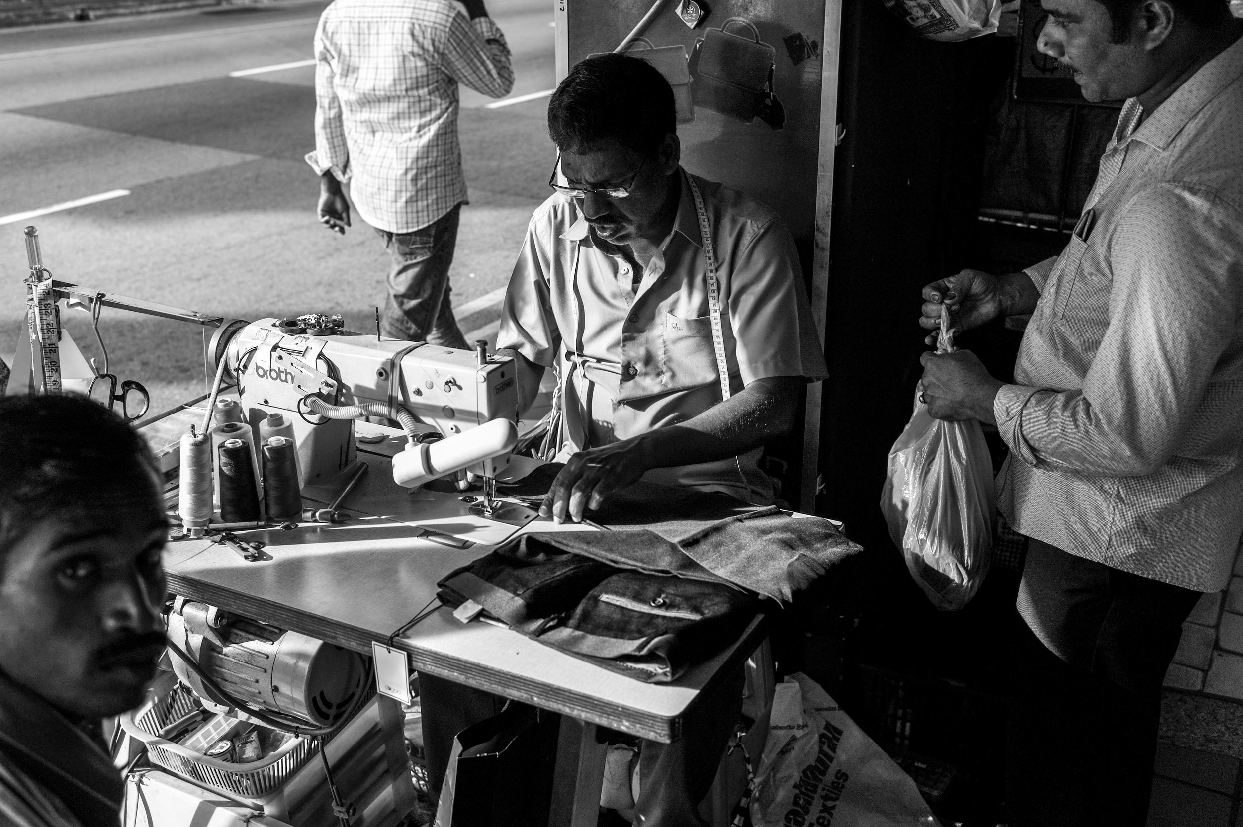   Services on a fast lane . A roadside tailor enjoys a relatively brisk business day as customers come forth to have their garments fixed. 