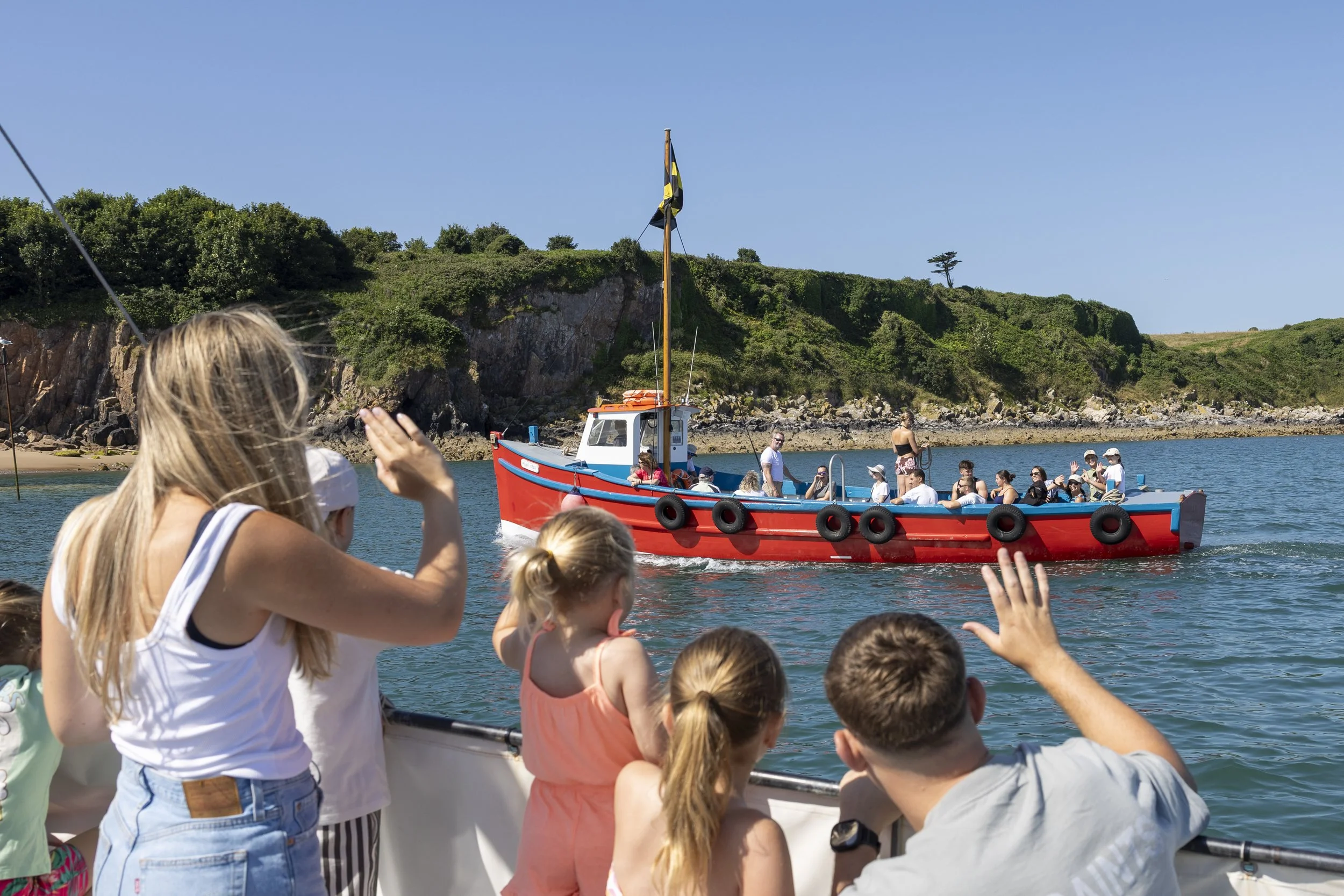 Group of people, including children and adults, on a boat watching another boat approaching with a rocky shoreline and green hills in the background.