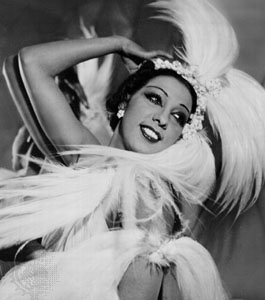 A black and white photo of Josephine Baker, a black woman with short black hair tucked into a large feather and crystal headpiece. She wears crystal earrings and bold makeup, posing and smiling into the distance.