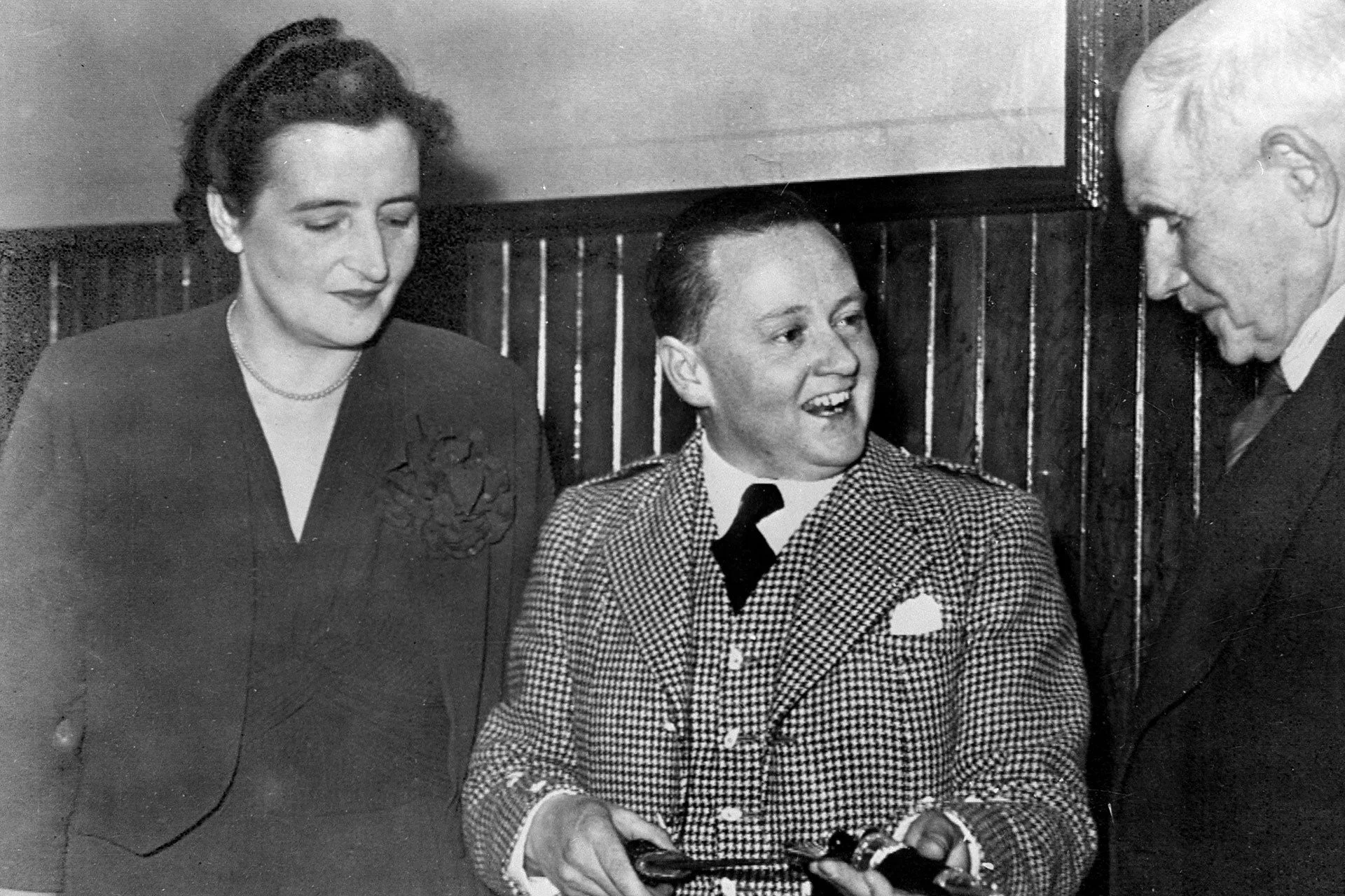 Sir Ewan Forbes, a white man with short dark hair, wears a suit and smiles at an older man. A woman stands with him.