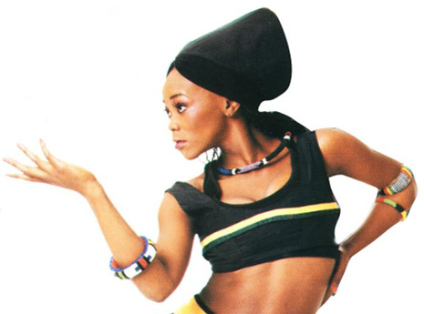 A black woman wearing a head wrap, a crop top, and multicolored bangles and necklaces poses with one hand on her hip and the other outstretched.
