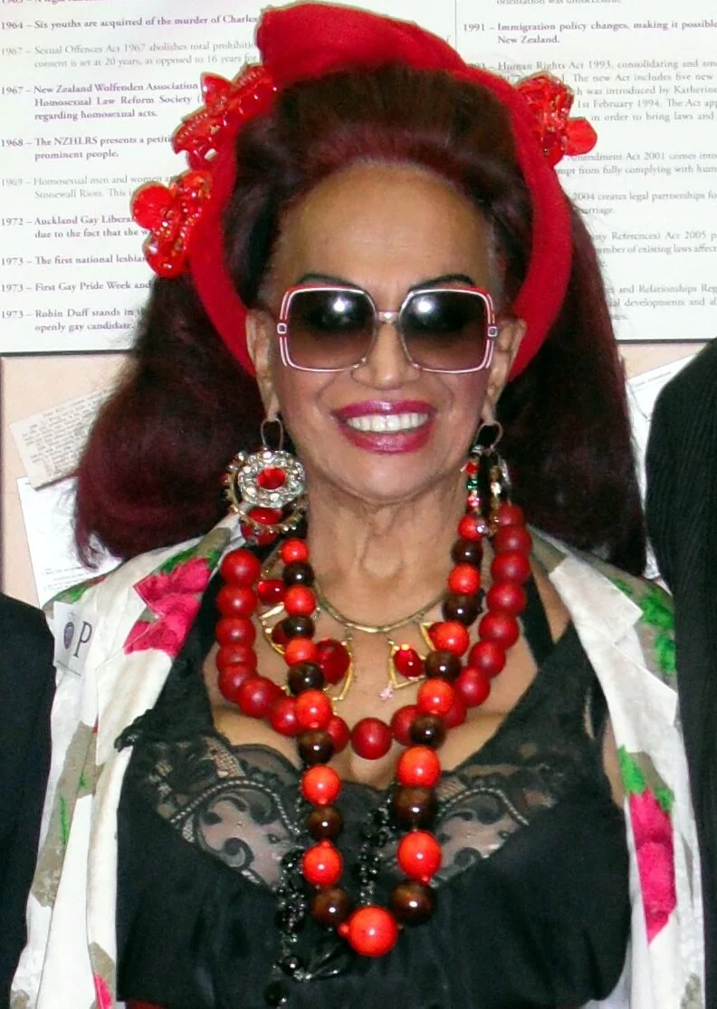 Carmen Rupe, a Māori woman with long brown hair wearing a red headpiece, red bead necklaces, red and white earrings, and sunglasses. She smiles.