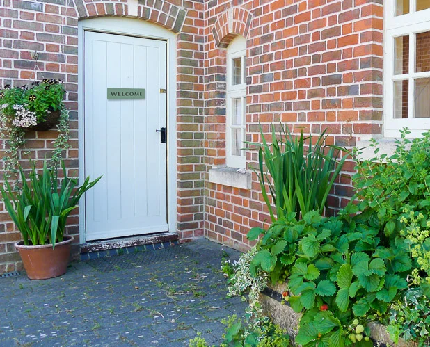 White door with a 'WELCOME' sign, surrounded by brick walls and green plants in pots on a paved walkway.