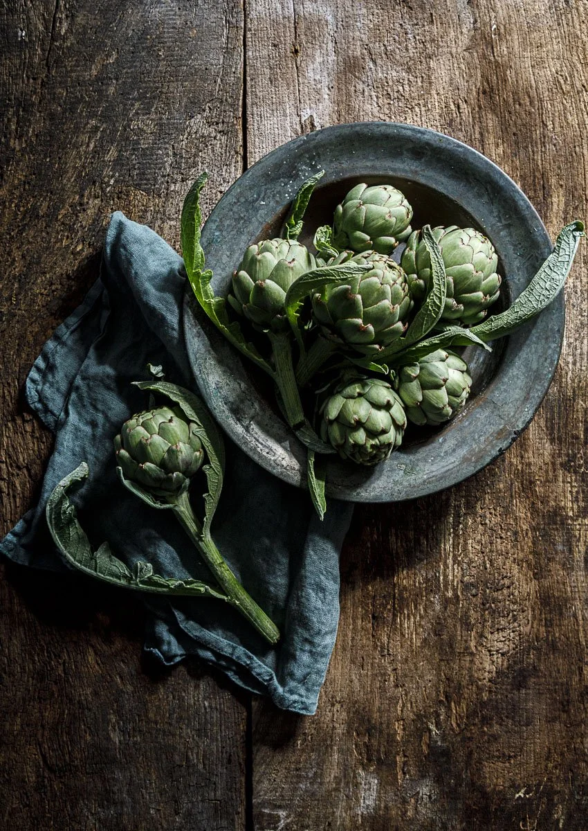 Fresh green artichokes on a rustic wooden table with some leaves and a blue cloth.