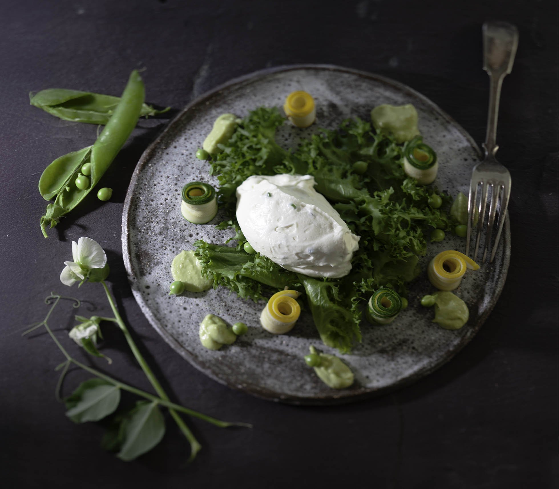 A plated salad with various green vegetables, a dollop of white sauce or cheese, and rolled yellow and green vegetable slices on a speckled ceramic plate, with an extra snap pea pod and a small white flower nearby.
