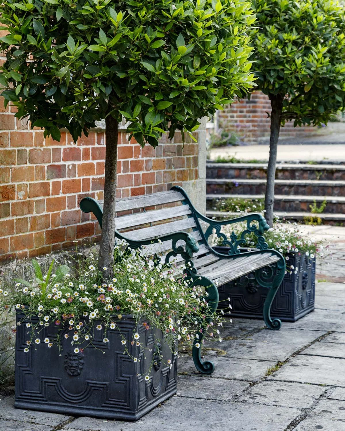 A bench with ornate metal armrests and backrest, painted green, situated on a stone pathway between two potted plants with daisies, with a brick wall and steps in the background.