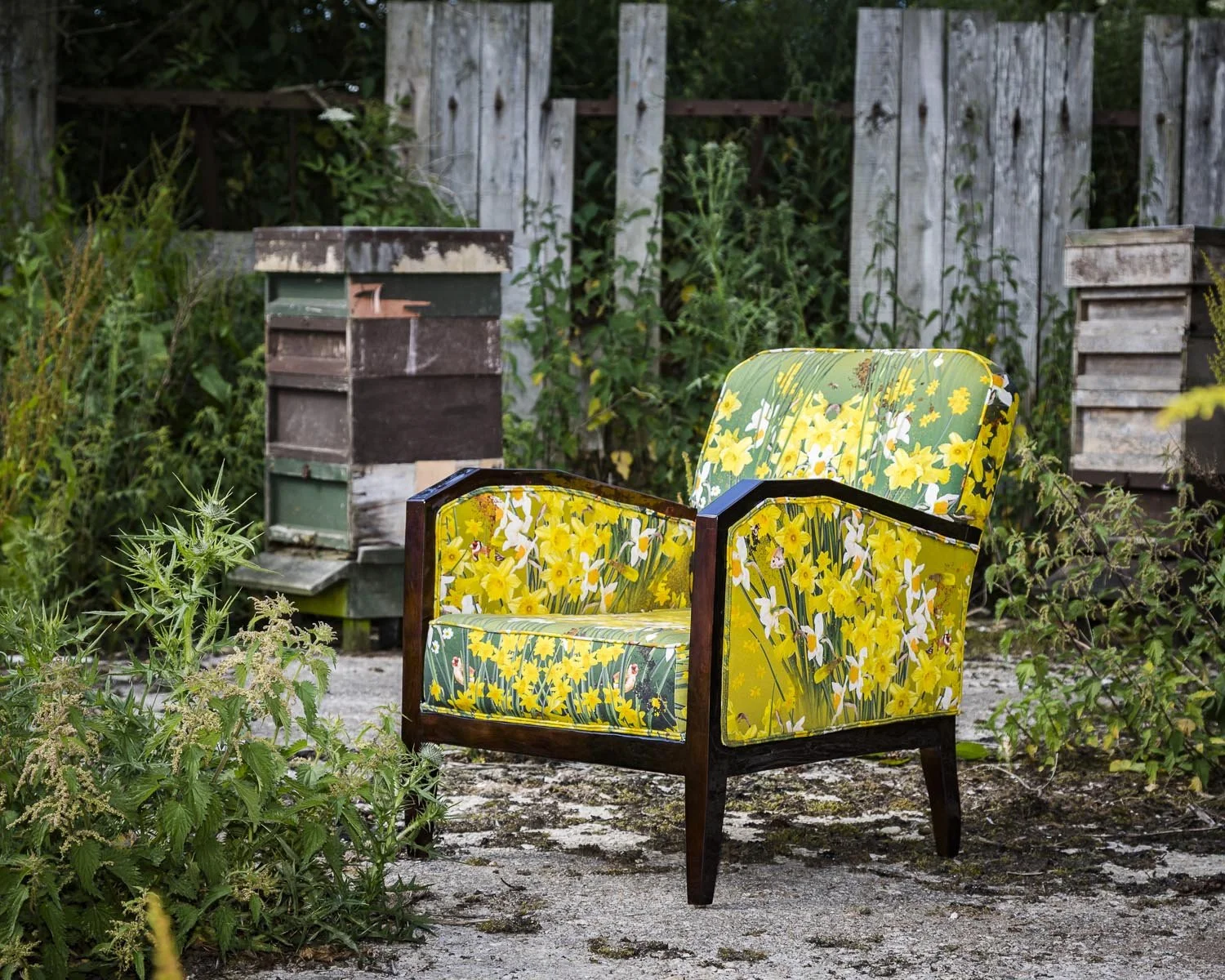 A yellow floral armchair with a dark wood frame outdoors near a weathered wooden fence and overgrown weeds, with a bee hive in the background.