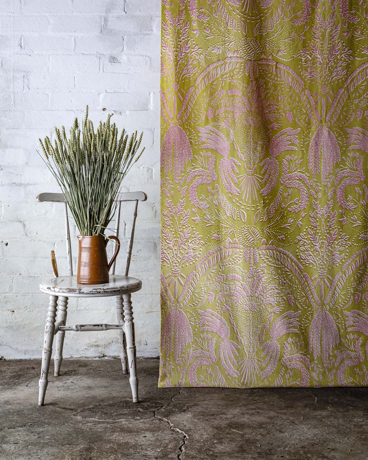A wooden chair with white paint chipping off, placed on a concrete floor against a white brick wall. A brown pitcher with green plants is on the chair. To the right, part of a pink and green patterned curtain is visible.