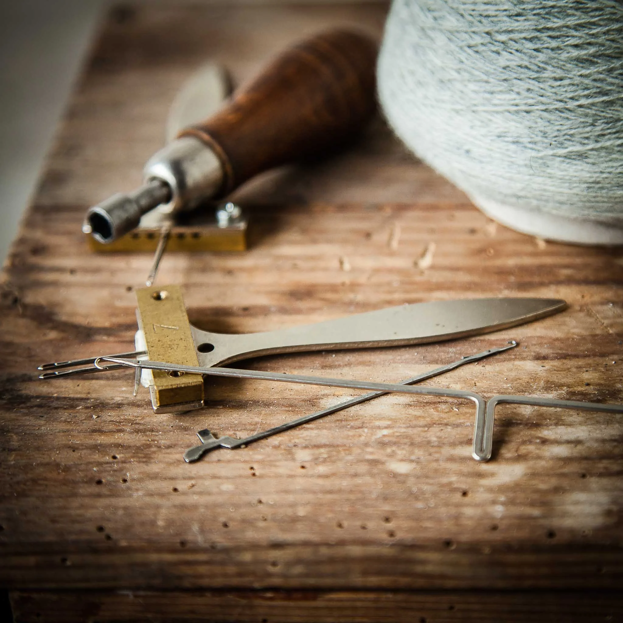 Tools including a draw gauge, a protractor, a needle, and a small hand tool are arranged on a wooden workbench with a spool of thread in the background.