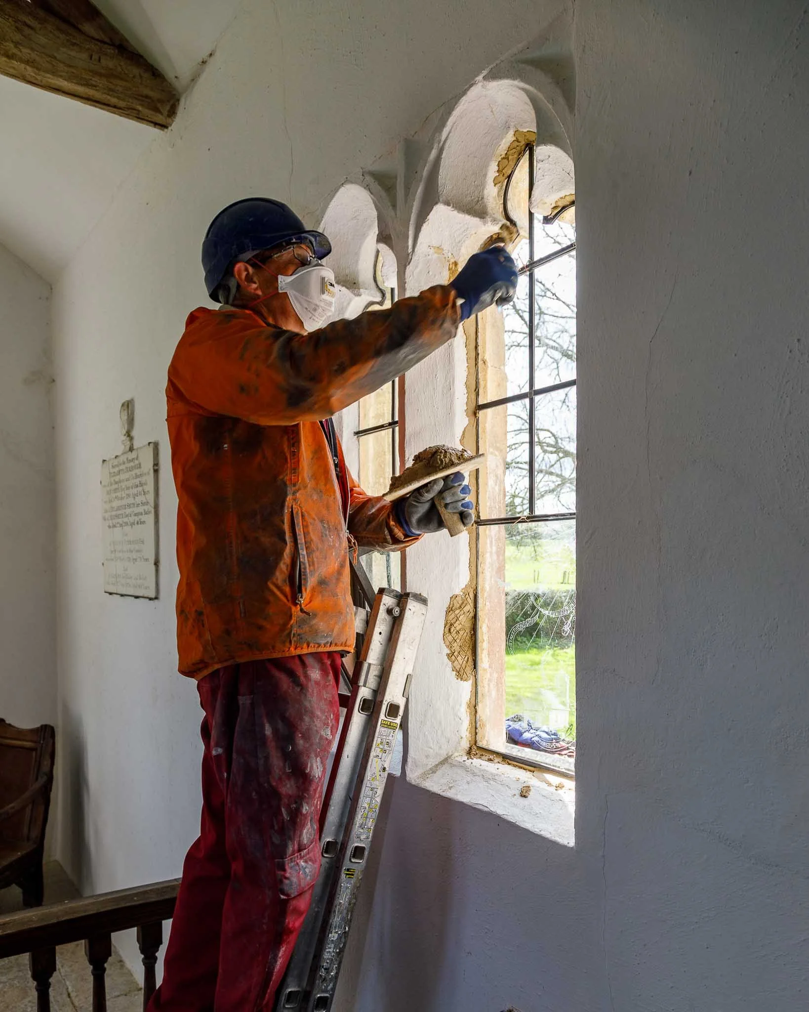 A worker wearing a helmet, mask, and gloves is repairing a leaded glass window in a church or historical building, standing on a ladder.