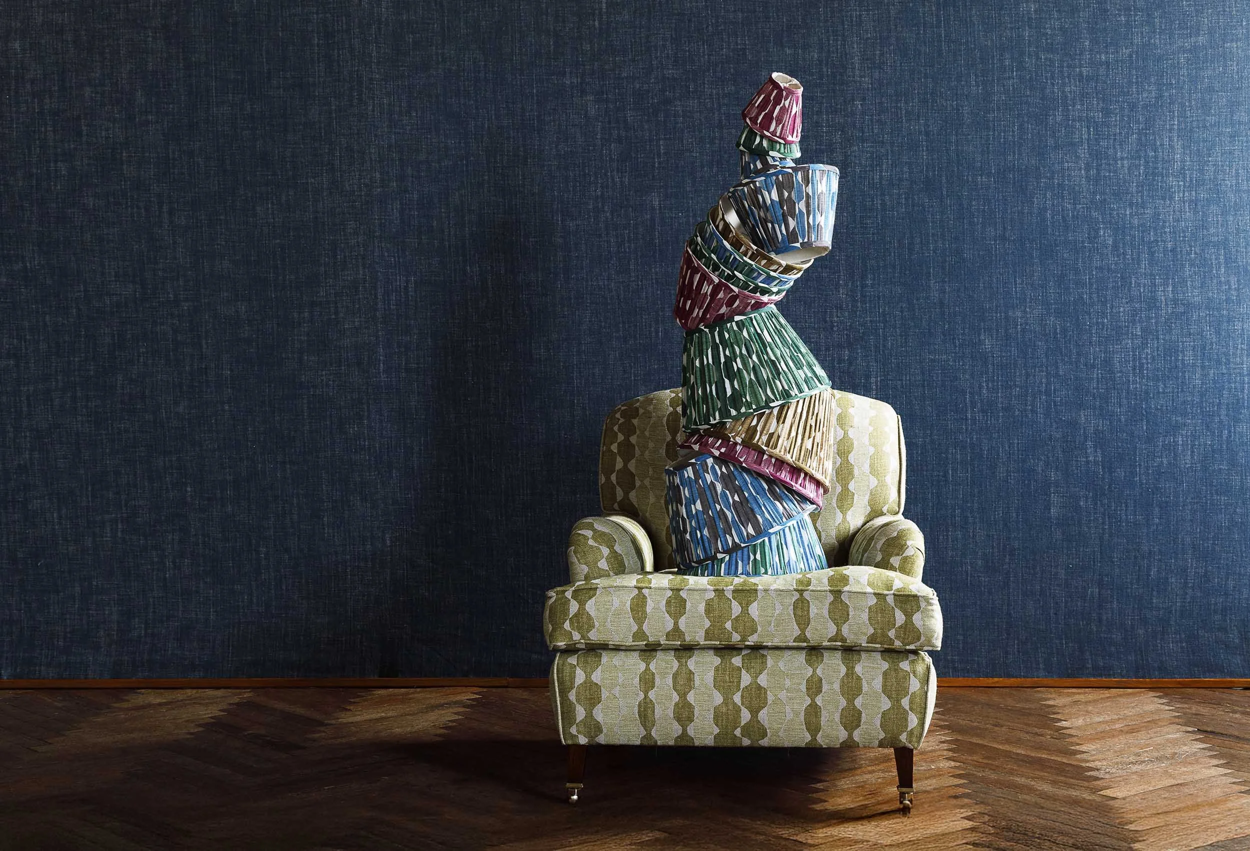 A green patterned armchair with a tall stack of colorful, patterned cupcake liners balanced on its back, against a dark blue textured wall and wooden floor.