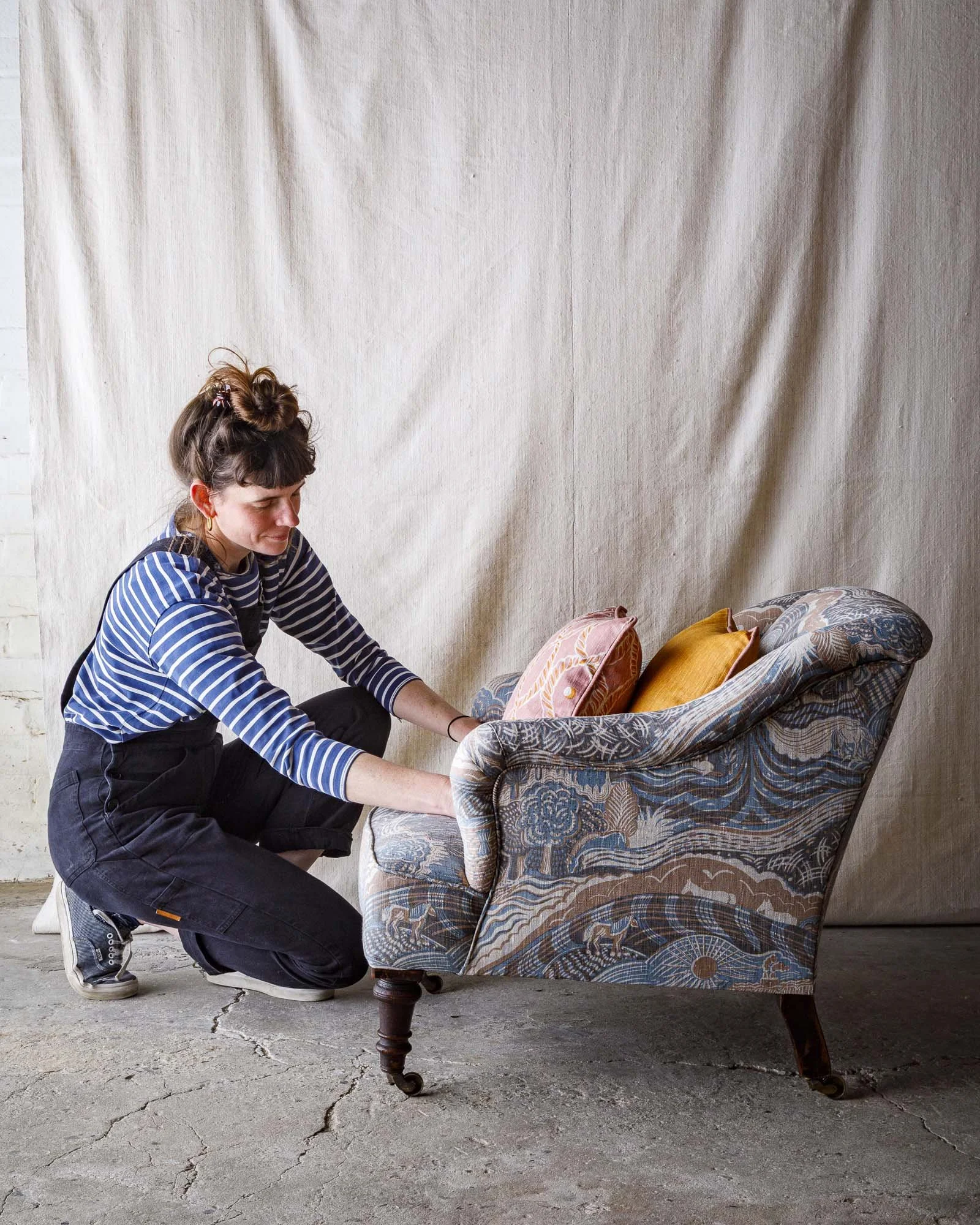 A woman with brown hair, wearing a striped shirt and black pants, kneeling on a concrete floor, adjusting a patterned vintage armchair decorated with colorful cushions in front of a beige backdrop.
