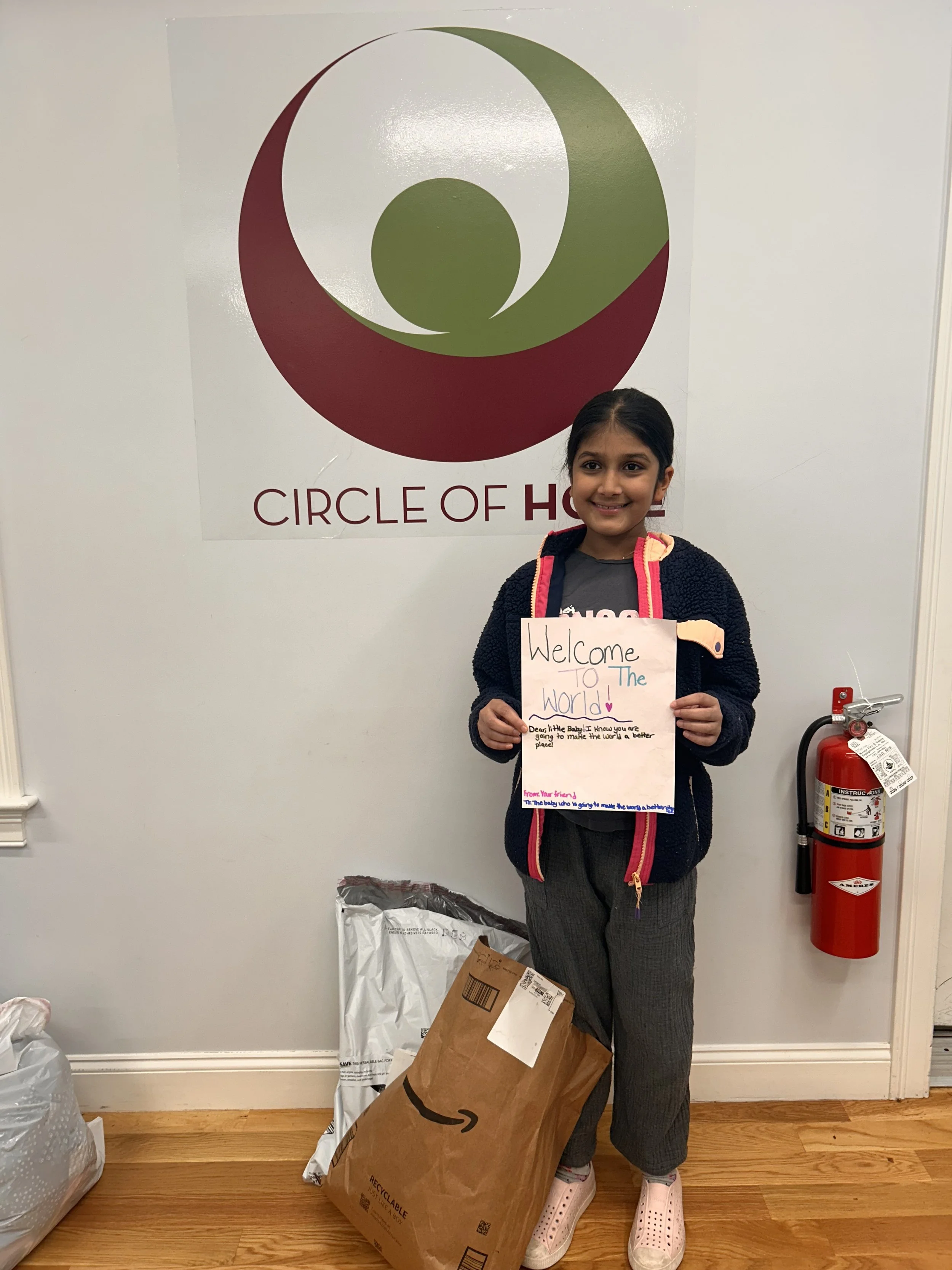 A young girl standing in front of the Circle of Hope logo holding a sign that says "Welcome to the World!"