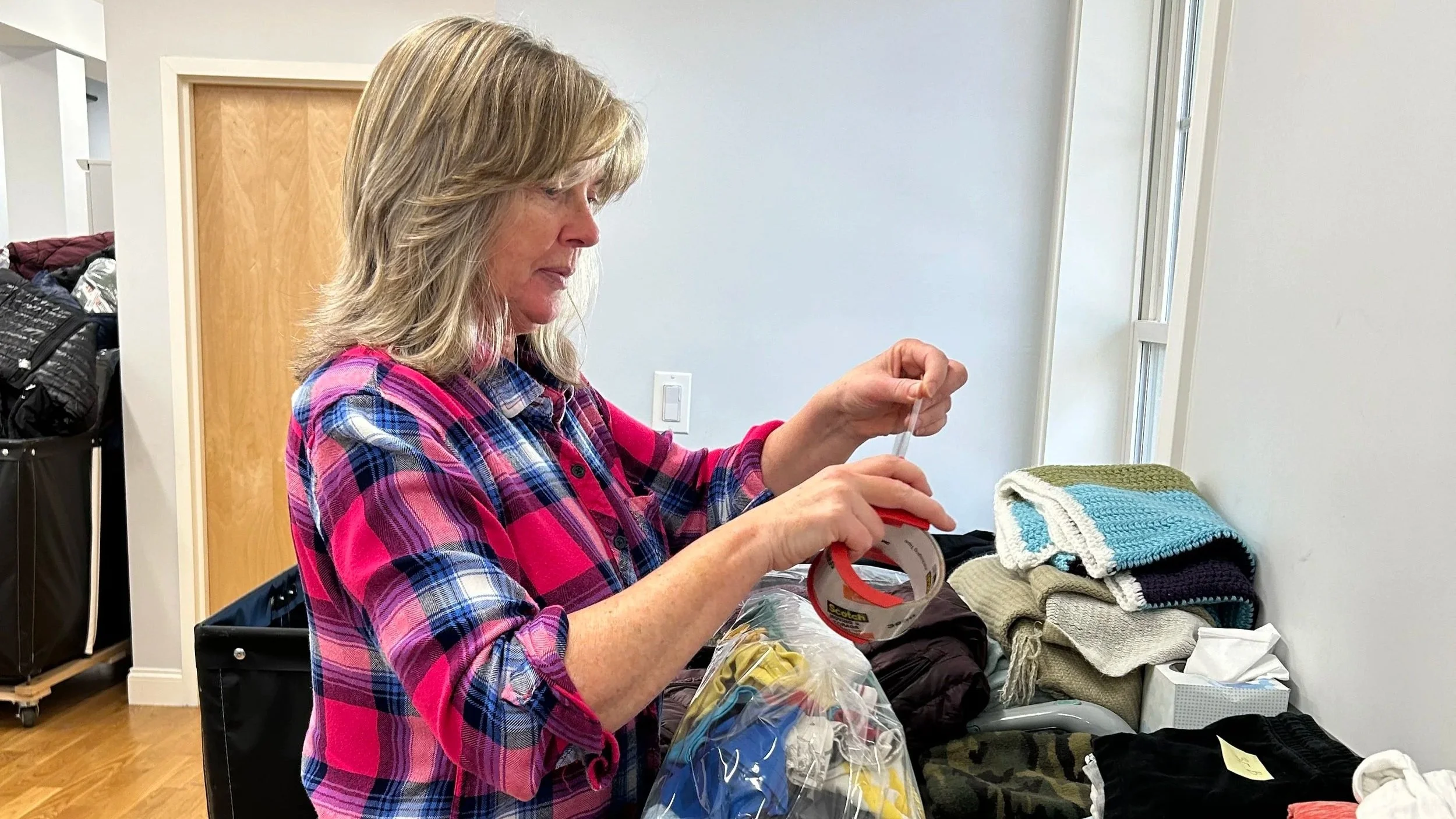 A woman standing in front of a counter full of clothing. She is taping up a bag of clothing donations.