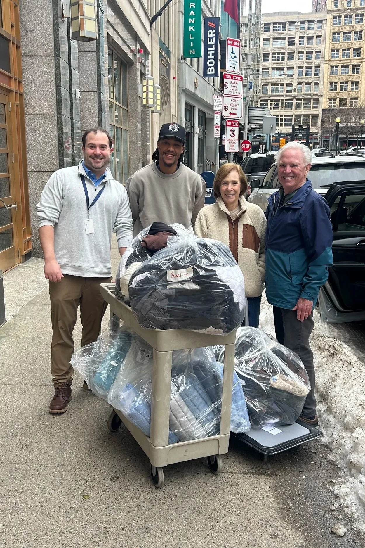 4 adults stand on a Boston sidewalk behind rolling carts full of bagged clothing donations.