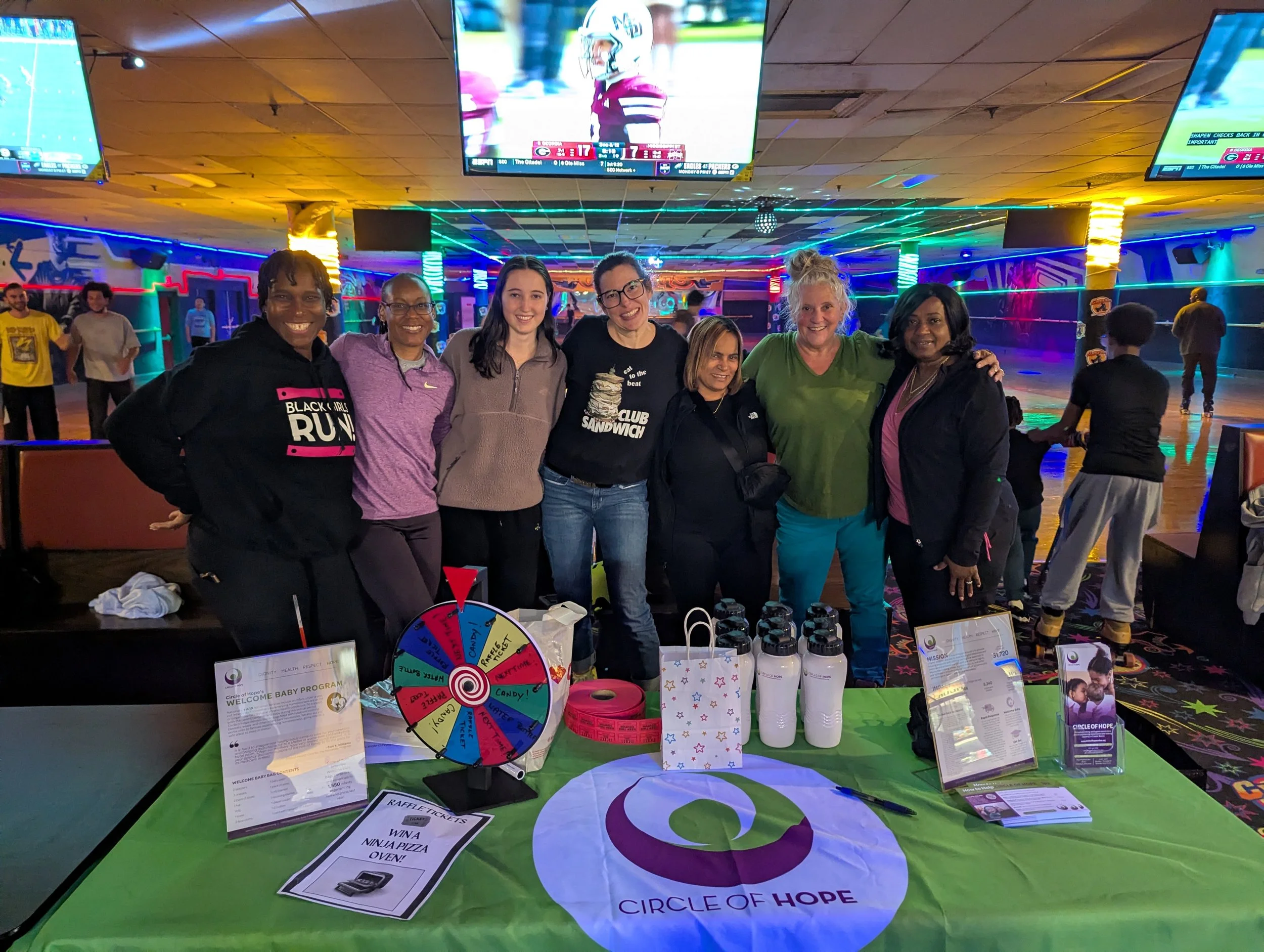 A group of 7 women stand behind a Circle of Hope table in front of a skate rink.