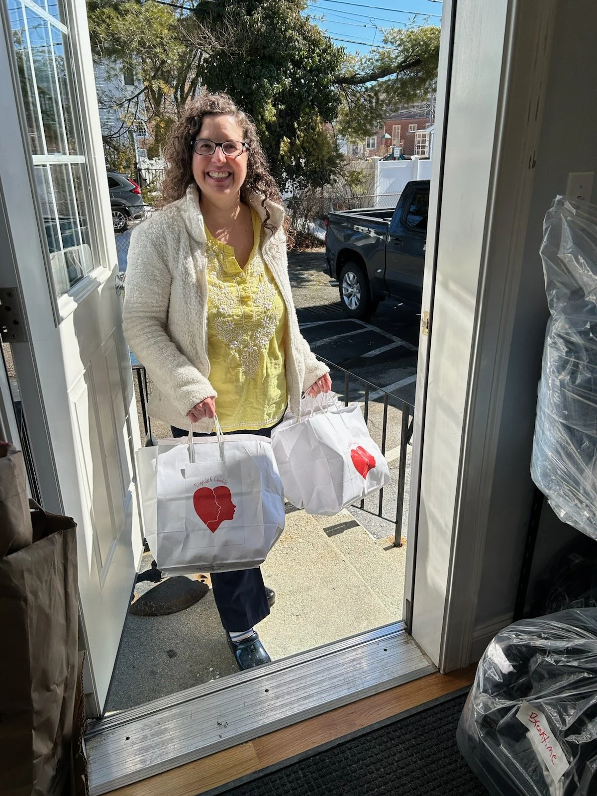 A smiling woman in a yellow shirt stands in the open doorway of Circle of Hope on a sunny day holding white bags full of toiletries.