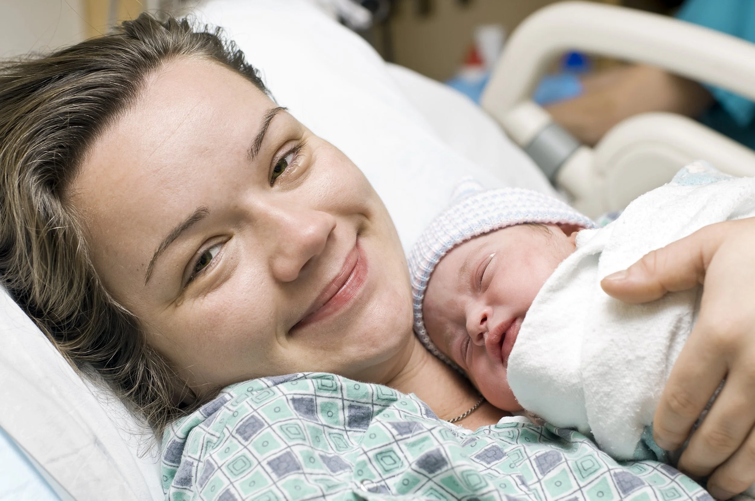 A mom laying in a hospital gown and bed holding her newborn wrapped in a blanket. Mom is smiling at the camera.